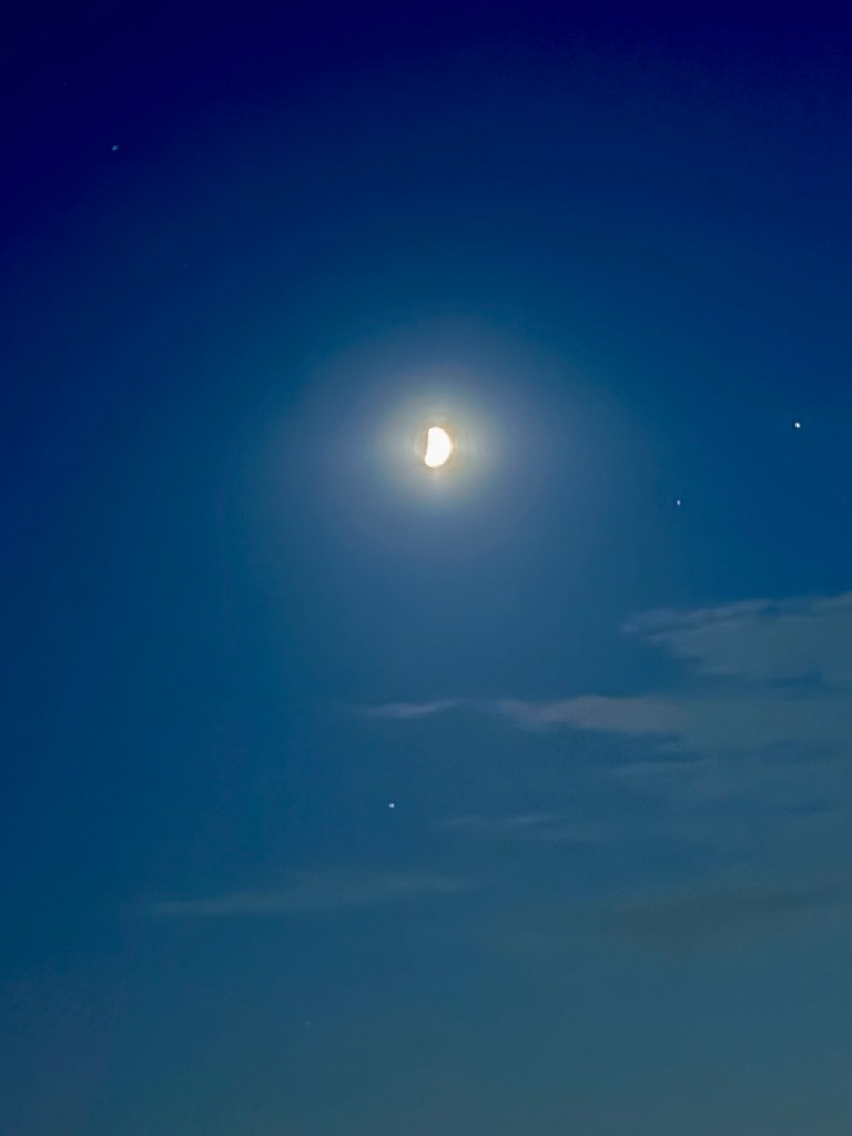 Bright Moon against a blue-gray evening sky, with several small points of light and faint wisps of cloud.