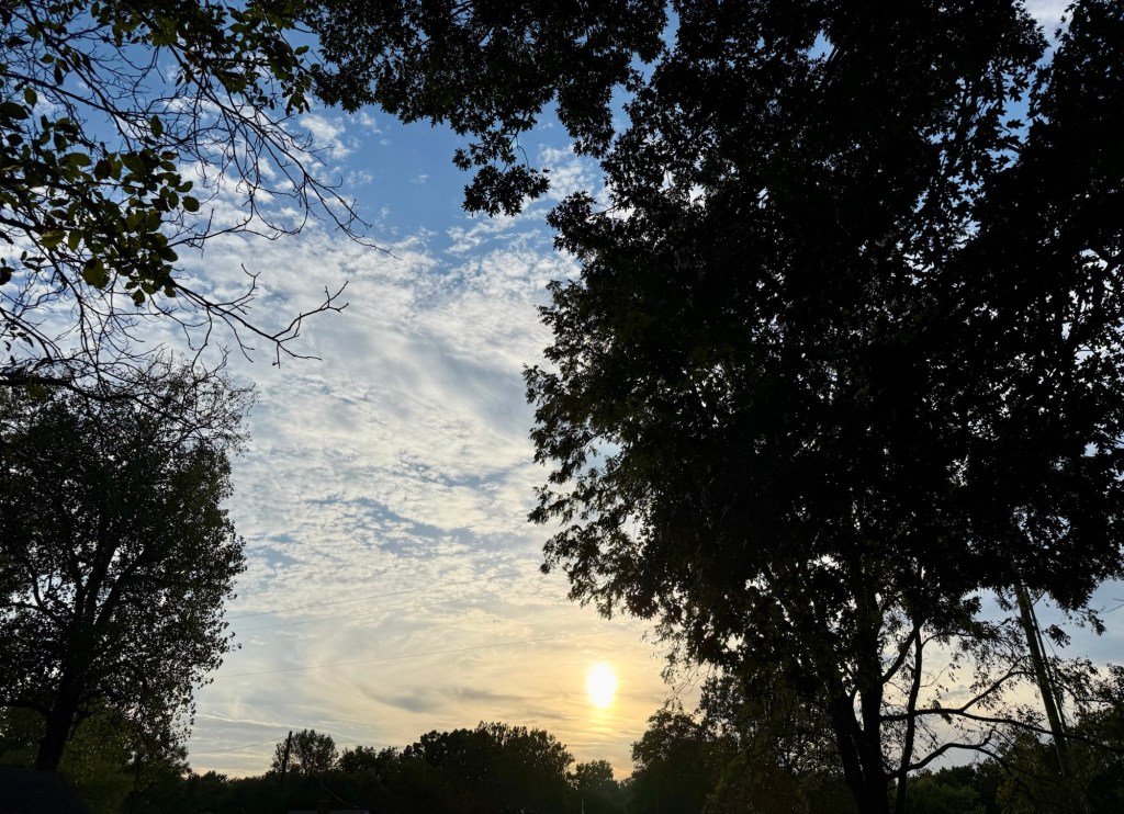 The lowering Sun glowing through patterned evening clouds framed by trees in Independence.