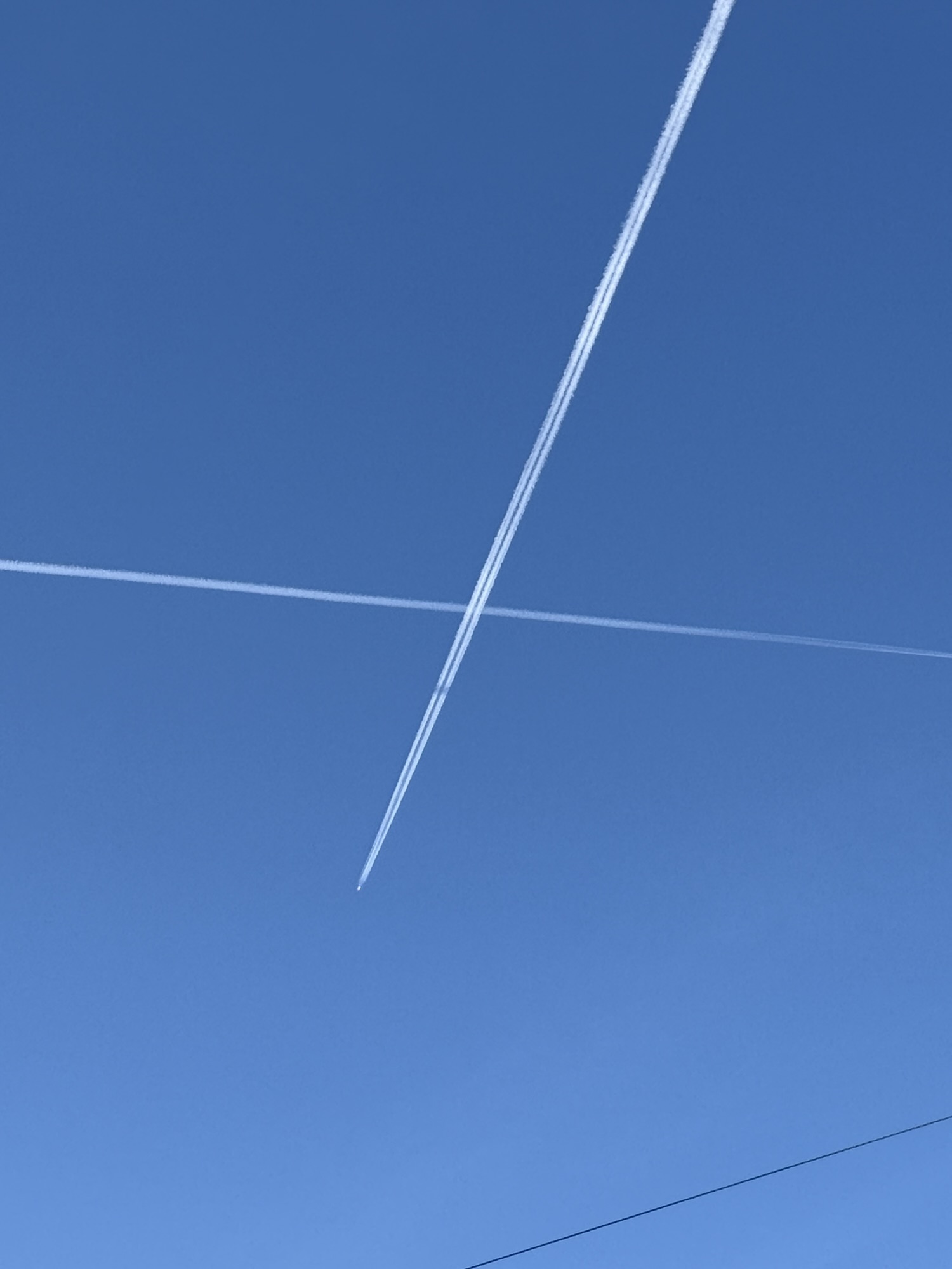 Two airplane contrails crossing in a blue sky, with an aircraft visible at the tip of the newer trail.