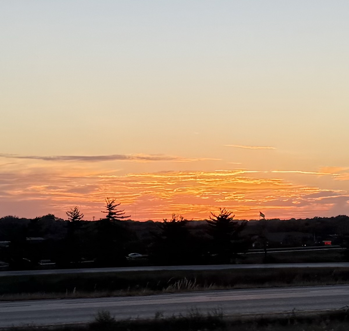 Orange and coral sunset illuminating layered clouds above a dark tree-lined horizon.