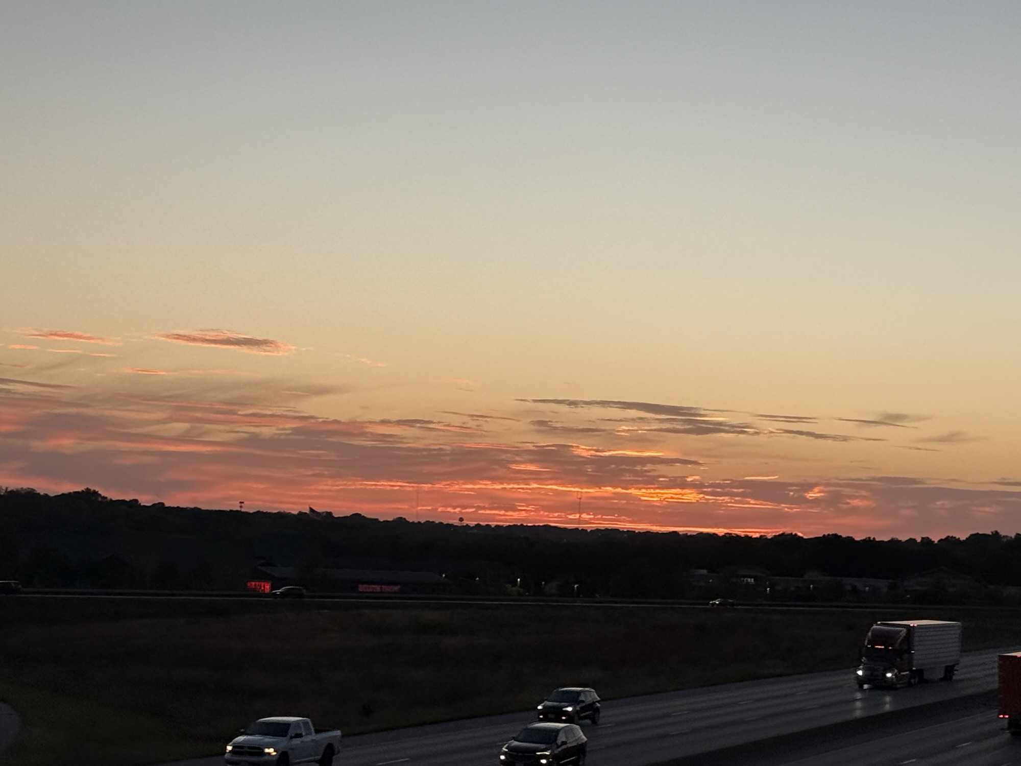 6:52 PM sunset with coral and peach clouds above the highway and passing traffic.
